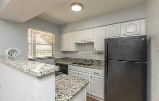 A modern kitchen featuring white cabinetry, a black refrigerator, and a granite countertop with gray and white speckles. There is a window with blinds allowing natural light, and a tiled backsplash complements the design. The space is clean and well-organized, creating a welcoming atmosphere.