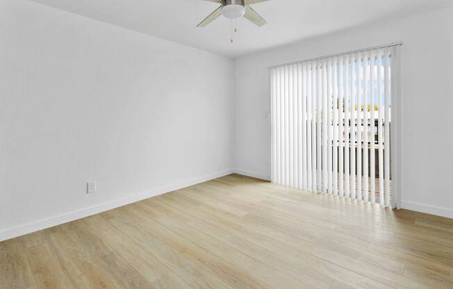 A room with a ceiling fan and light fixture, wooden flooring, and a window with blinds.
