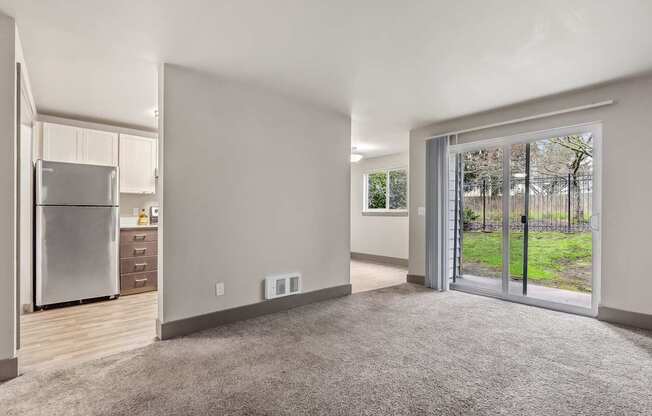 A spacious, empty room with a grey carpet and a sliding glass door leading to a green outdoor area.