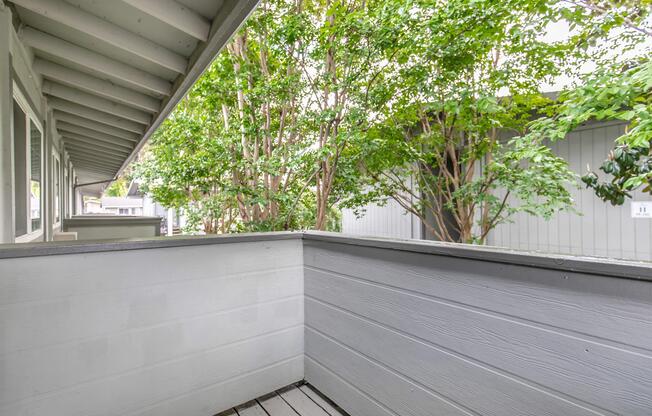 View of a balcony with a wooden railing, surrounded by lush green trees. The space features a light-colored wooden floor and walls. Natural light filters through the trees, creating a serene outdoor atmosphere. No people or furniture are visible in the scene.
