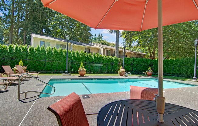 A shaded poolside seating area here at Asbury Park featuring a round outdoor table with chairs beneath a large umbrella, overlooking a clear rectangular swimming pool with metal handrails, surrounding lounge chairs, potted planters, and a fenced perimeter framed by tall evergreen hedges and mature trees in a peaceful community setting.