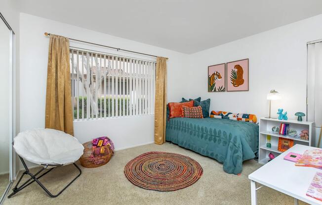 A cozy bedroom featuring a light blue bedspread with decorative pillows, a circular woven rug, and a small white desk with colorful items. There are two framed artworks on the wall, a large window with vertical blinds, and a comfortable chair in the corner. Warm curtains complement the bright and inviting space.
