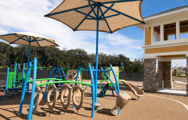 A playground with a blue and yellow swing set and a tan umbrella.