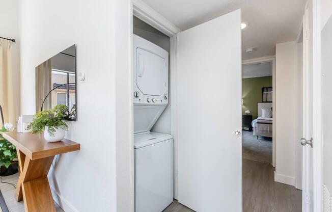 A compact laundry area featuring a stacked washer and dryer, located behind a door to the left of a modern living space. A minimalist table with a decorative plant sits nearby, and a mirror reflects the bright, light-colored walls. In the background, a glimpse of a bedroom is visible.