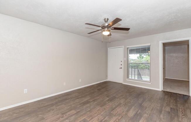 A spacious living room featuring a ceiling fan, light-colored walls, and wooden flooring. There is a front door with a window next to it, and a doorway to another room on the right. Natural light streams in through the sliding glass door, which leads to a balcony or patio area outside.