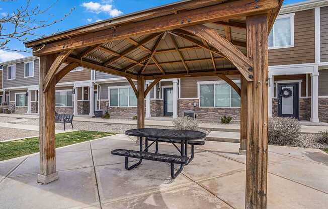 A wooden pergola with a black picnic table is in front of a building.