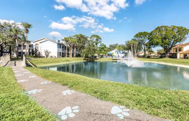 a pathway leading to a pond with a fountain and houses in the background