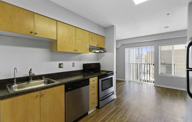a kitchen with wooden cabinets and stainless steel appliances with a view into the spacious living area with plank flooring and sliding glass doors at Illumina Apartment Homes, Seattle, WA