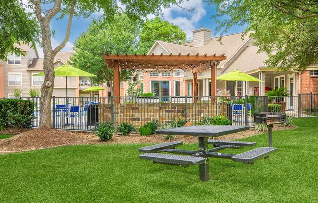 A landscaped outdoor area featuring a picnic table on green grass, surrounded by various plants. There is a stone patio with a pergola in the background, along with bright green umbrellas over blue chairs. The setting is part of an apartment complex, with residential buildings visible nearby.