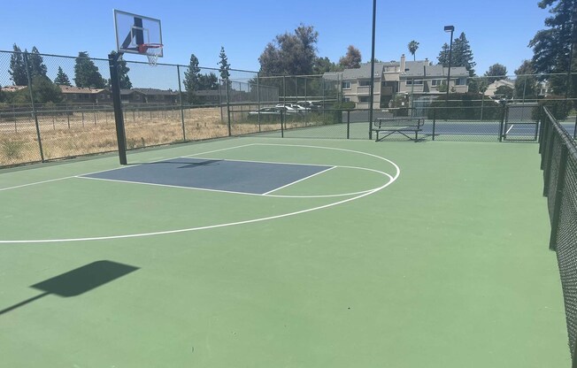 A view of an empty outdoor basketball court with a green surface, a single hoop, and a three-point line. In the background, there's a fenced area and a glimpse of residential buildings and trees under a clear blue sky.