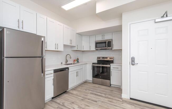 Modern kitchen featuring stainless steel appliances including a refrigerator, dishwasher, and oven. The cabinets are white with sleek hardware. A microwave is mounted above the stove. The floor is wood-look laminate, and there is a white front door visible on the right.