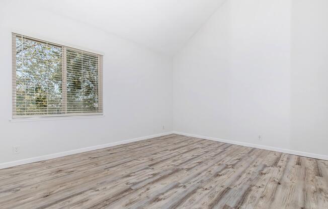 Empty room with white walls, a high ceiling, and a large window letting in natural light. The floor is covered with light wood laminate, and there are no furnishings present. The window features blinds, and outside greenery is visible through the glass.