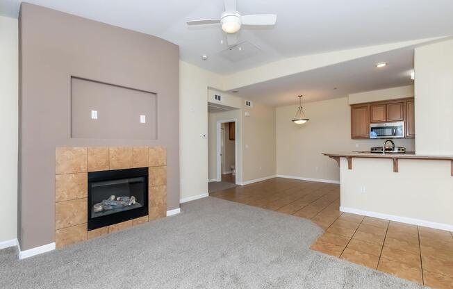 Cozy living area featuring a carpeted floor, a decorative fireplace with a tile surround, and an open layout leading to a kitchen with wooden cabinets. A ceiling fan is installed, and there is natural light from the adjoining space, enhancing the inviting atmosphere.