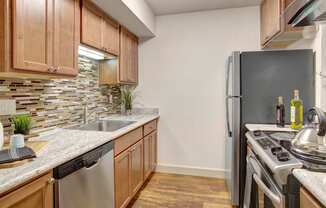 A modern kitchen here at Heritage Hills showcasing warm wood cabinetry, sleek countertops, and a mosaic tile backsplash that adds visual interest. The space features a stainless-style sink, dishwasher, refrigerator, and range, along with wood-look flooring, under-cabinet lighting, and a streamlined galley layout designed for efficient everyday cooking and entertaining.