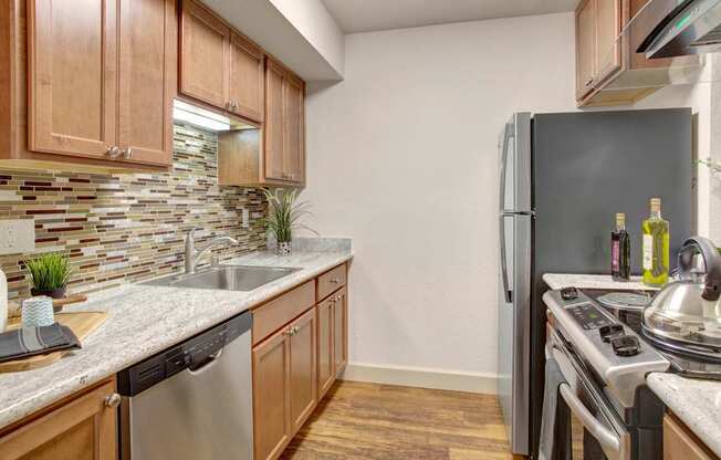 A modern kitchen here at Heritage Hills showcasing warm wood cabinetry, sleek countertops, and a mosaic tile backsplash that adds visual interest. The space features a stainless-style sink, dishwasher, refrigerator, and range, along with wood-look flooring, under-cabinet lighting, and a streamlined galley layout designed for efficient everyday cooking and entertaining.