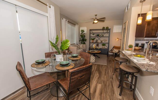 A modern dining area with a round glass table set for four, featuring dark wooden chairs. The space includes a cozy living room in the background with a large bookshelf, decorative plants, and natural light from the windows. The kitchen bar is visible, adorned with fruit and casual decor.