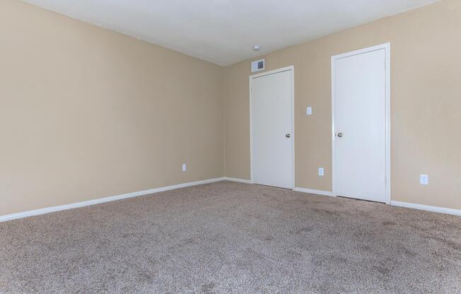 A simple, empty room with light beige walls and plush, beige carpet. The space features two closed white doors on the right, one likely a closet, and a small white vent above. Natural light filters in, giving the room a bright and inviting feel, though it remains uncluttered and minimalistic.