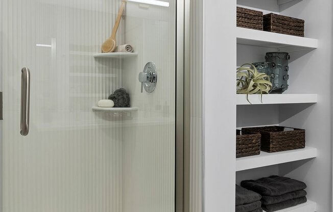 A white bathroom with a glass shower door and shelves with towels and other bathroom accessories.