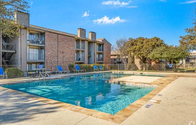 A swimming pool in front of a brick building with blue chairs around it.