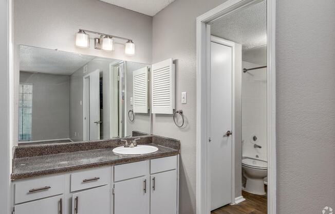 A modern bathroom featuring a double-sink vanity with a granite countertop, a large mirror with three light fixtures above, and a bathroom entrance leading to a toilet. The walls are painted in a light gray color, and there is a framed window visible in the background.