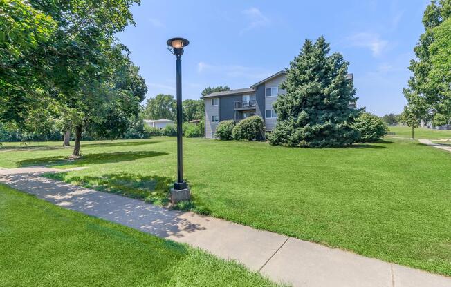A well-maintained grassy area with a paved walkway leads to a multi-story residential building. The scene is framed by lush trees, and a lamppost adds to the inviting atmosphere under a clear blue sky.