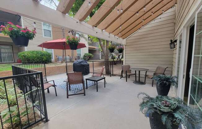 A covered outdoor patio here at Trinity Place with a concrete surface and comfortable seating arranged for conversation. A wooden pergola overhead is accented with string lights and hanging planters, creating a welcoming atmosphere. A grilling area and surrounding greenery add to the relaxed community setting.