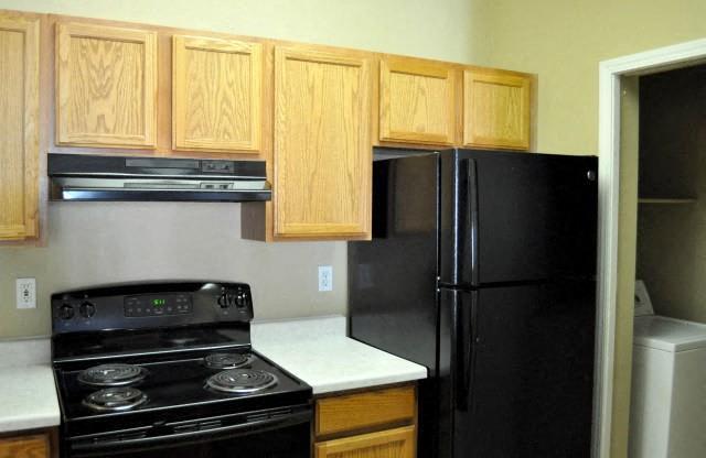 North Creek Apartments - Kitchen with Black Electric Appliances and Light Colored Wooden Cabinets with Access to Laundry Closet Area.