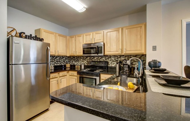 U-shaped kitchen with black countertops, stainless steel refrigerator, microwave and stove. Ample cabinet space with blonde cabinets above and below counter with multi-colored tile backsplash