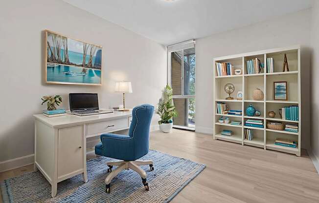 A room with a blue chair, a desk with a laptop, a bookshelf with books, and a painting on the wall.