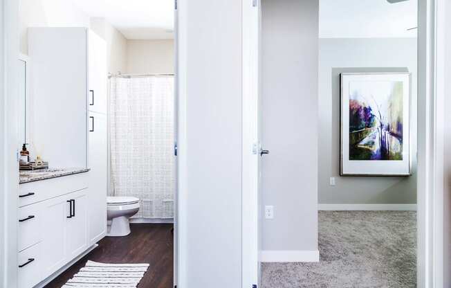 Modern apartment bathroom featuring stain-resistant floors and elegant granite countertops. The adjacent spare bedroom boasts plush carpeting and serene nature views.