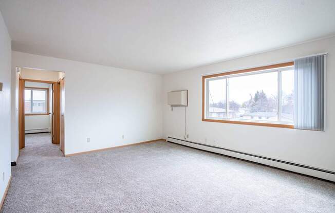 A large, empty room with a carpeted floor and a window with curtains. West Fargo, ND Saddlebrook Apartments