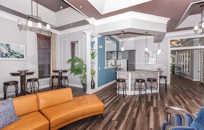 Modern lounge area featuring a brown leather sofa, bar stools, and a kitchen island. The room boasts large windows allowing natural light, stylish decor, and a mix of dark wooden flooring and light wall colors, creating a bright and inviting atmosphere.