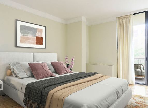 A bed with a white headboard and a grey blanket is in the foreground of a room with a framed picture on the wall and a chair by the window.
