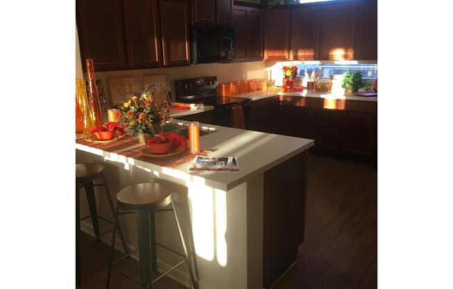 A beautifully lit kitchen featuring wooden cabinets, a modern stove, and a countertop with decorative bowls of fruit. The dining area has two bar stools, and the warm sunlight casts shadows across the space. A floral centerpiece and vibrant orange accents add a cozy, inviting atmosphere.