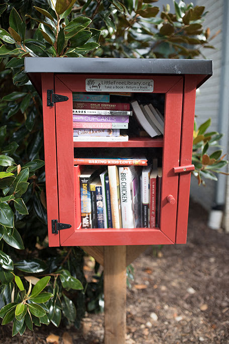 A red Little Free Library box is filled with books and sits in front of a green bush.