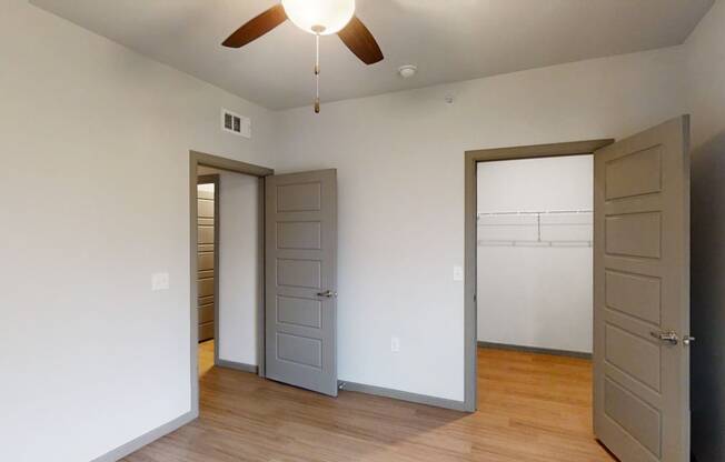 A minimal bedroom here at 49 West Apartments featuring light gray walls, a wooden floor, two open doors, and a ceiling fan with lights. Bright, serene, and spacious ambiance.