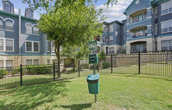 A tree in front of a building with a green trash can in front of it.