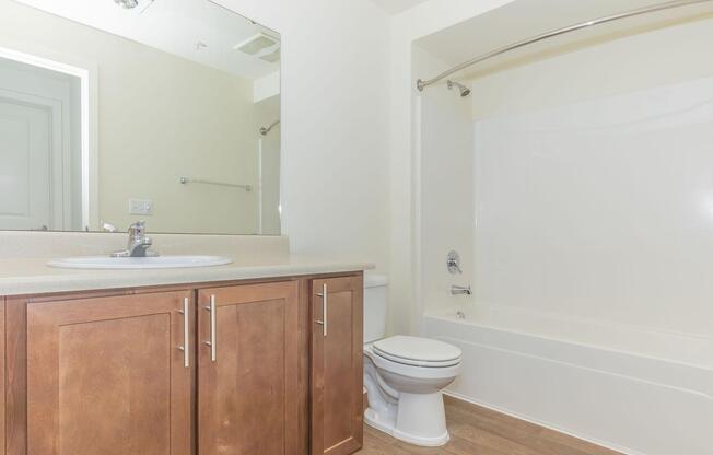 A clean, modern bathroom featuring a beige countertop with a sink, a mirror above, a white bathtub with a shower rod, and a toilet. The walls are light-colored, and the flooring is wooden, creating a bright and spacious atmosphere.