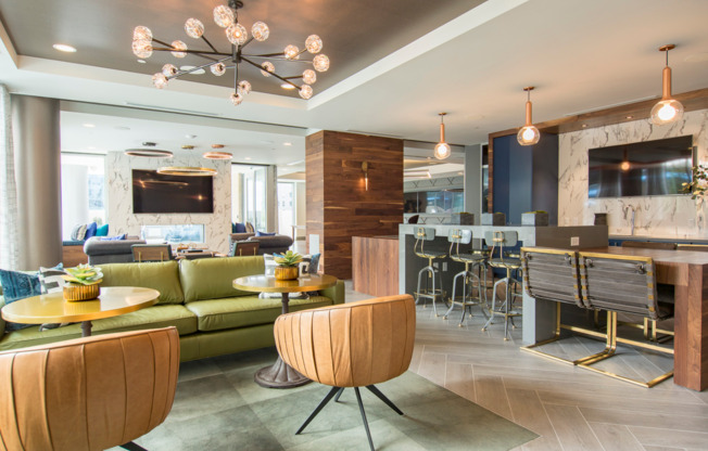 A bright lounge space with large windows, colorful leather couches and armchairs, a bar with an HDTV, a see-thru fireplace, wood-paneled walls, and modern light fixtures.
