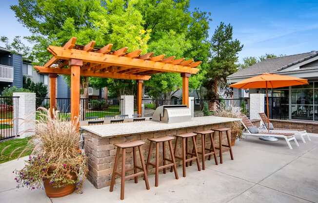 A wooden pergola with a bar and stools is surrounded by greenery.