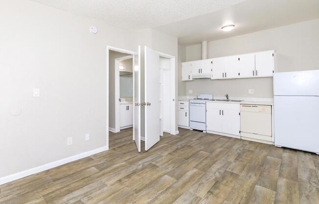 A small, modern kitchen and living area featuring wood-effect flooring, white cabinetry, a refrigerator, and a stove. Two open doors show a bathroom and a closet. The walls are light-colored, and the space is bright and airy, suggesting a recently renovated apartment.
