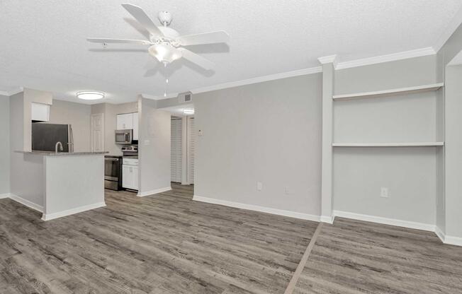 Bright, open living area featuring a ceiling fan and light wood-like flooring. The kitchen is visible in the background with stainless steel appliances and a breakfast bar. Light gray walls provide a modern feel, with built-in shelves on one side enhancing the space's functionality.