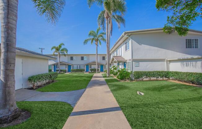 A sunny residential complex featuring well-maintained pathways, manicured lawns, and palm trees. The two-story buildings are surrounded by greenery, and the clear blue sky adds to the bright atmosphere. The scene conveys a peaceful and inviting environment for residents.