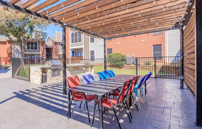 A covered outdoor patio area featuring a large table with red and blue chairs, surrounded by a fence. In the background, residential buildings and a grassy area are visible. Strings of lights hang from the wooden pergola, providing a cozy ambiance for gatherings. A grill is located at one end of the patio.