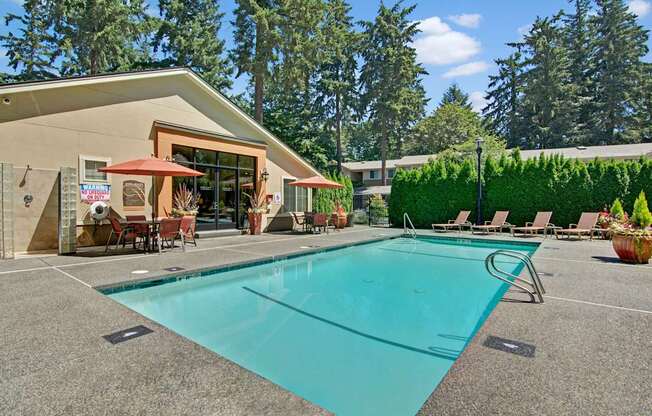 A sunlit outdoor swimming pool here at Asbury Park featuring a clear rectangular pool with metal handrails, a wide textured pool deck, lounge chairs along the edge, shaded patio tables with umbrellas, and a clubhouse building with large windows, all surrounded by tall evergreens and manicured hedges creating a relaxed community setting.