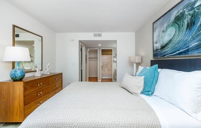 A modern bedroom featuring a king-sized bed with light gray bedding and turquoise accent pillows. There is a wooden dresser with a decorative lamp and a figurine. A large mirror reflects the space, and a wave-themed artwork hangs above the bed. The room has a bright, airy feel with neutral colors and natural light.