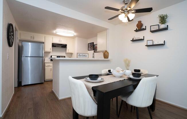 Modern kitchen and dining area featuring a black dining table with white chairs, set for a meal. The kitchen includes stainless steel appliances, a white countertop, and decorative items on the shelves. A ceiling fan provides light, and the space is bright and inviting with neutral colors.