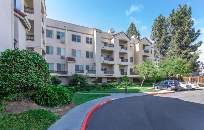A multi-story residential building with a beige exterior, featuring balconies and landscaped greenery, including shrubs and trees. A paved driveway curves around the property, with a parked car visible. The scene is set under a clear blue sky.
