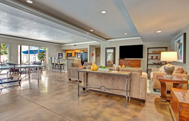 A game and lounge room here at Bridges with tan sofas, wood accent tables, ping pong table, bar-height kitchen counter with stools, TV wall, and polished concrete floors in a bright, open layout.