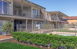 A view of a two-story apartment community featuring balconies, a fenced area, and manicured hedges. In the foreground, well-maintained shrubs line a grassy area, while the background shows a swimming pool surrounded by a safety fence. Clear blue skies above enhance the inviting atmosphere.
